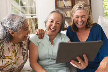 Diverse senior female friends laughing at home, one holding tablet