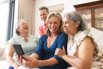 Diverse senior female friends laughing at home, looking at tablet
