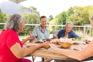 Diverse senior female friends enjoying meal outdoors, saying grace and praying