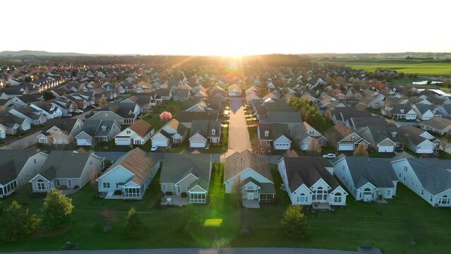 Suburban neighborhood at sunset, with rows of similar houses, green lawns, and a street leading directly into the glowing sun on the horizon. Aerial truck shot.