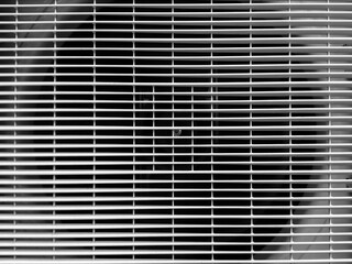 Household air conditioner with gray protective grill. It is blowing cool air into room. Grill is made of plastic and has holes to prevent objects from being sucked into air conditioner. Close-up.