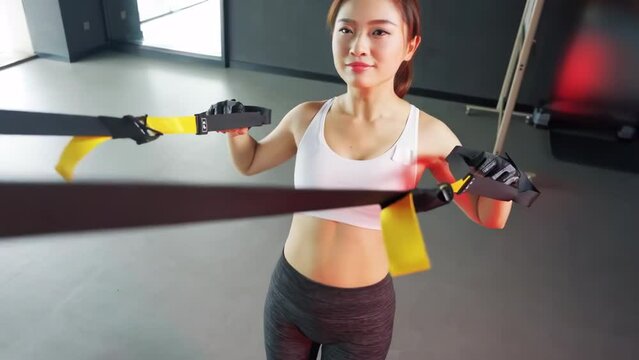 Athletic muscular fitness woman showing suspension training, performing pull-ups exercise with trx straps at gym. Lifting up and down to maintaining muscles