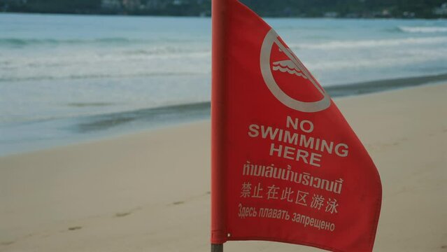 Red warning flag indicating text No Swimming Here in multiple languages placed on beach with waves. multi-language message inform beachgoers of water hazard. Safety notice or warning sign on beach