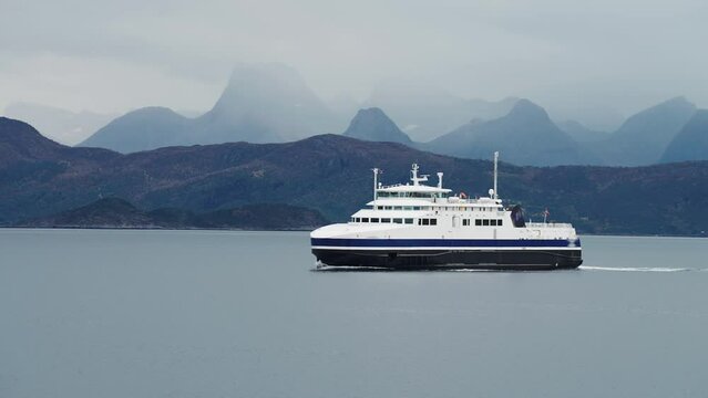 A huge passenger ferry crosses Norwegian fjod. mountains wrapped in fog tower on the horizon.