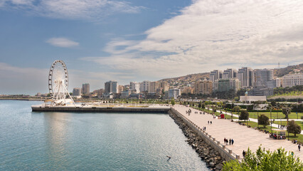 People stroll along the waterfront promenade near the Baku Eye ferris wheel in Baku, Azerbaijan, with modern buildings lining the background under a partly cloudy sky
