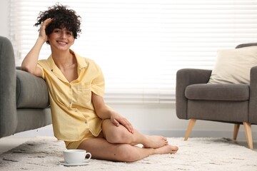 Beautiful young woman in stylish pyjama with cup of drink on floor at home