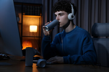 Young man with energy drink and headphones playing video game at wooden desk indoors