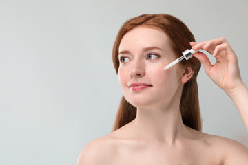 Beautiful woman with freckles applying cosmetic serum onto her face against grey background. Space for text