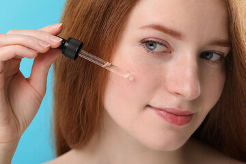 Fototapeta premium Beautiful woman with freckles applying cosmetic serum onto her face against light blue background, closeup