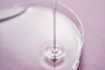 Serum or cosmetic oil flows into a transparent bowl on a purple background.