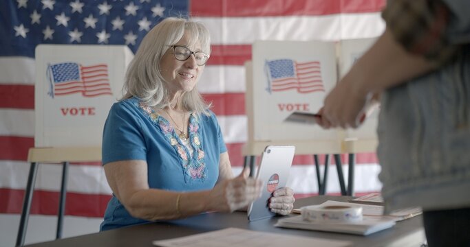 Three quarter angle, medium shot, elderly woman election worker checks voter identification for young female voter using tablet computer at polling station. Flag and voting booths behind