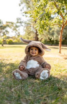 A little girl with a bunny onesie in a park siting down on the grass.