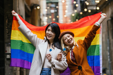 Happy Asian lesbian couple smiling while holding a lgbt rainbow flag outdoors.