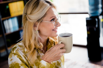Photo portrait of lovely mature woman hold drink coffee look empty space dressed stylish shirt office day light home interior workspace