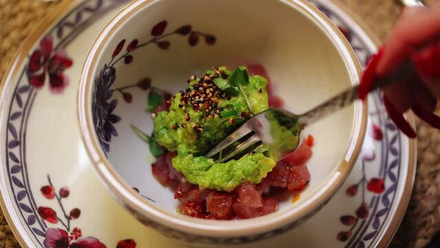Unrecognizable female with long red nails holding fork and trying first piece of tuna tartare with spicy guacamole sauce. Experienced food critic visiting freshly opened restaurant for review.