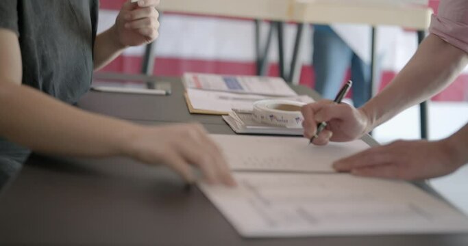Medium shot, profile, young, red-haired woman, poll worker at voting center, checks identification for male voter. He signs in and she starts handing him a ballot. Voting booths and US flag behind