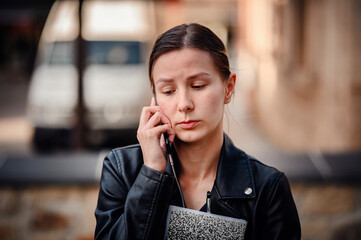 A young woman in a black leather jacket holds a notebook and talks on her phone, appearing focused and serious in an urban setting.