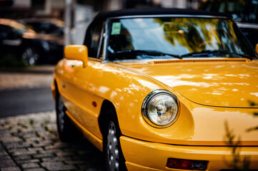 Close-up of a yellow classic car parked on a cobblestone street, with plants and another car in the background. The vintage vehicle's design and vibrant color stand out.