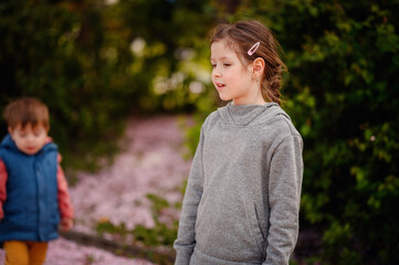 Young girl and her little brother playing outdoors on a spring day, surrounded by fallen cherry blossom petals and lush greenery.