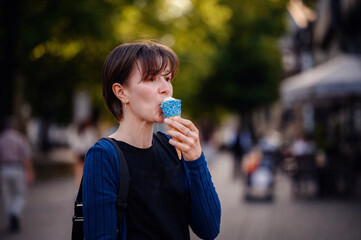 A woman with short hair enjoys an ice cream cone decorated with blue sprinkles on a sunny day in town. The background shows a lively street scene with blurred people and greenery.