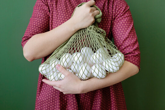 Person Holding a Mesh Bag Full of Eggs Against Green Background