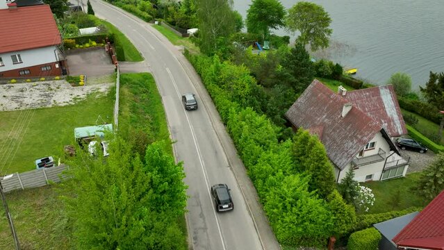 An aerial view of a road alongside a lake, with houses and green foliage lining the sides with cars on the road.