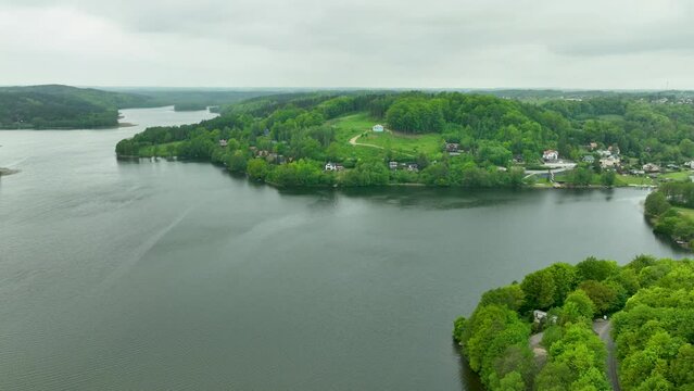 An aerial panorama of a lakeside village with houses nestled among green hills and trees, reflecting in the water.