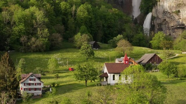 A picturesque Swiss village with traditional houses and a charming church sits at the foot of Seerenbach Falls. The lush greenery and towering cliff create a serene backdrop.