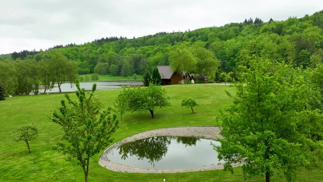 A serene park landscape with a small pond, trees, and a cozy cabin in the background, surrounded by green hills