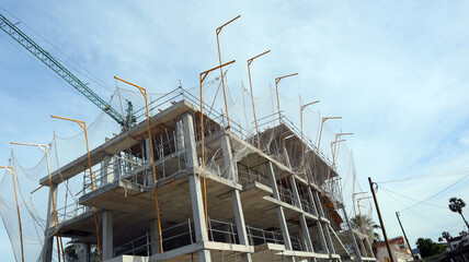 Multi-story concrete building is under construction, featuring scaffolding and safety nets. Project is located outdoors on clear land with a blue sky overhead.