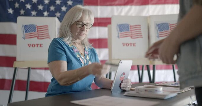 Three quarter angle, medium shot, elderly woman election worker checks voter identification for young female voter using tablet computer at polling station. Flag and voting booths behind