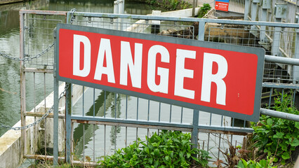 Close up of large danger sign in red with bold white font on metal fence overlooking canal water