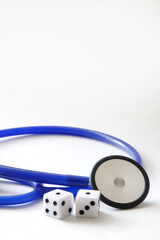 a blue stethoscope next to two dice in front of a white background