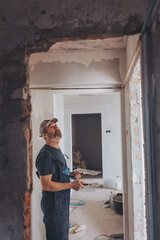 Side view of a plasterer standing in a room in renovation process and using tools for plasterwork and skim coating walls. A worker is renovating house and doing manual building works.