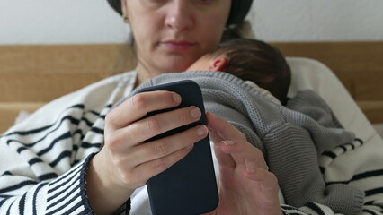 Close-up of mother holding cellphone device while holding newborn baby resting on chest, laid in...