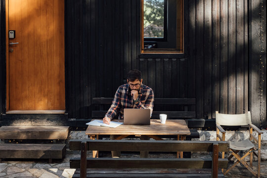 Handsome Man Sitting in Front of Mountain Cabin and Working on Laptop