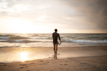 Male Surfer Walking Towards the Waves at a Sandy Beach During Sunset