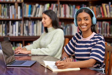 Portrait, university and woman in library with headphones, laptop or notes with bookshelf. Smile, book and girl with research for scholarship, study and streaming music at table for college education