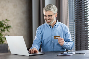 Mature man using credit card on laptop for online shopping in bright modern office