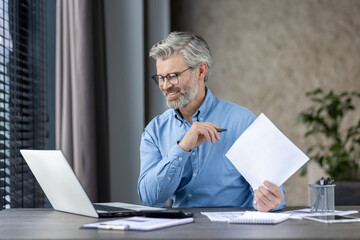A middleaged man is smiling while working on a laptop at his home office, holding papers