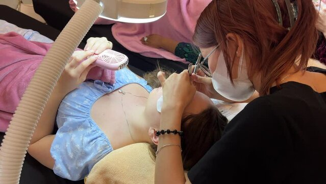 Video of process of eyelash extension, young lashmaker sitting over client lying on couch and gluing fake lashes on lid using tweezers. Concentrated master holding tools and touching cilia.