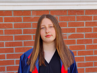 The image shows a young girl with long, straight hair standing in front of a red brick wall. She is wearing a high school graduation dress. The girl is not happy and looks directly at the camera.