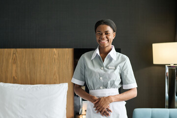 Waist up portrait of young Black woman as housekeeper wearing uniform and smiling at camera...