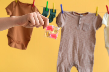 Female hand with gel capsule for washing and clothes hanging on clothesline against yellow...