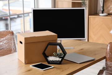 Desk with tablet computer, laptop, PC monitor and cardboard box in office on moving day