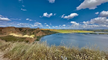 A panoramic view of the Barrancas de Burujón, Spain. Showcasing spectacular cliffs, a serene river and lush fields under a vibrant blue sky with scattered clouds.