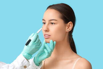 Doctor in protective gloves drawing marks on woman's nose for plastic surgery against blue...