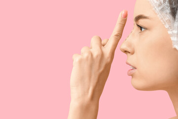 Young woman in medical hat checking her nose before plastic surgery against pink background, closeup