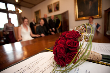 Red rose wedding bouquet on table in registry office, wedding ceremony attendees blurred in the...