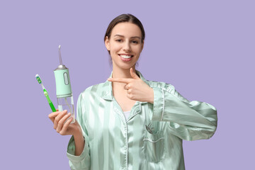 Beautiful young woman pointing at oral irrigator and toothbrush on lilac background